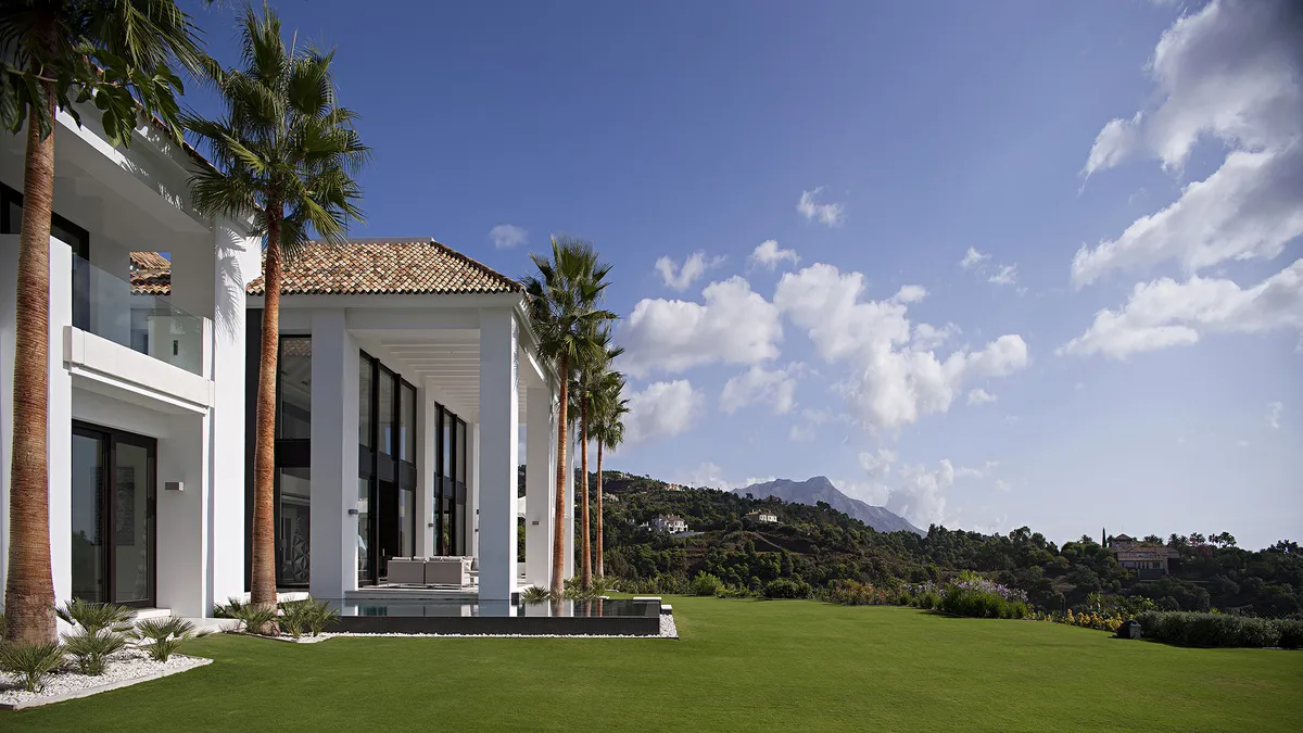 Villa Palo Alto's modern white exterior, palm trees, infinity pool, and lush green lawn against Marbella mountains.