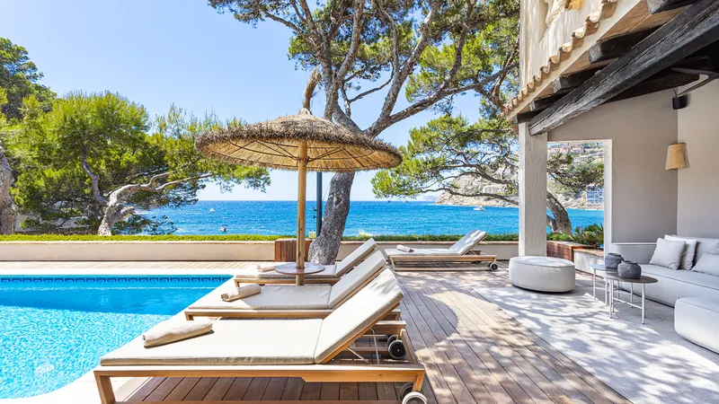 Villa Mañana — Poolside lounge with sea view Villa Mañana's poolside lounge area with sun loungers, a straw umbrella, and the Mediterranean Sea in Mallorca.