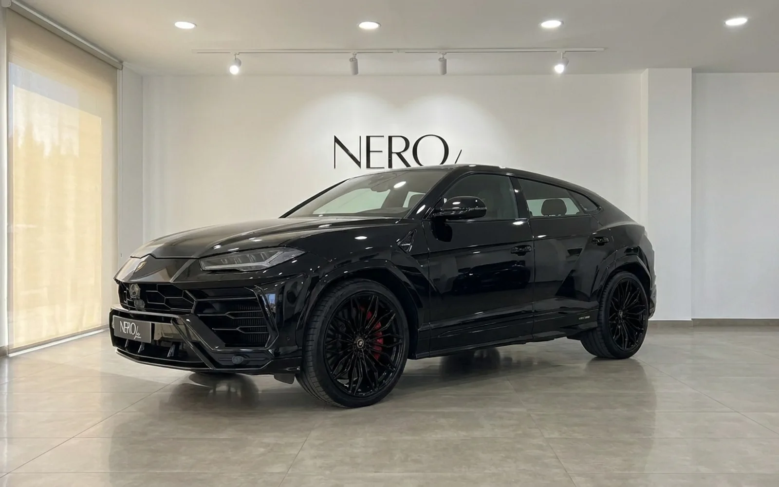 The gloss black Lamborghini Urus SE is elegantly displayed from a front three-quarter angle inside a modern showroom in Barcelona.