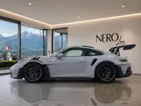 The gloss Arctic Grey Porsche GT3 RS is parked indoors, showcasing its side profile and large rear wing, with a stunning mountain view of Zurich in the background.