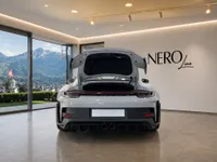 The Porsche GT3 RS in gloss Arctic Grey is shown from the rear with its frunk open, parked in a modern showroom overlooking Zurich's mountains.