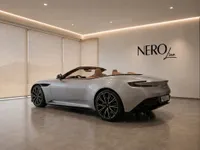 The metallic silver Aston Martin DB12 Volante, with its top down and tan interior, is showcased from a rear three-quarter angle in a modern showroom in Lausanne.