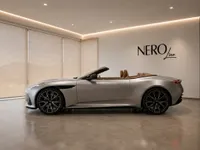 The metallic silver Aston Martin DB12 Volante, with its tan leather interior, is elegantly displayed in a modern showroom in Lausanne.