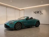 The metallic teal Aston Martin Vantage V8 Roadster is showcased in a modern showroom with its convertible top down.