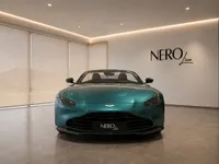 The metallic teal Aston Martin Vantage V8 Roadster is prominently displayed from the front in a showroom in Lausanne.