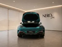 The rear view of a metallic teal Aston Martin Vantage V8 Roadster with its trunk open, displayed in a showroom in Lausanne.