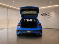 The blue Audi RS3 Performance Edition is seen from the rear with its trunk open, showcasing its spacious cargo area in a Zurich showroom.