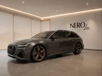 The gloss dark metallic grey Audi RS6 Avant Performance is showcased in a modern showroom in Lausanne, viewed from the front three-quarter angle.
