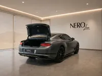 The Satin Anthracite Grey Bentley Continental GT is shown from the rear with its trunk open, parked in a modern showroom in Lausanne.