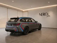 The matte grey 2023 BMW M3 Competition Touring with M-stripes is parked in a modern showroom in Zurich.