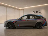 The matte grey BMW M3 Competition Touring with distinctive M stripes is showcased in a side profile view within a modern showroom in Zurich.