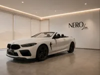 The white BMW M8 Competition Cabriolet is showcased in a modern showroom in Lausanne, featuring its sleek front three-quarter view with the top down.