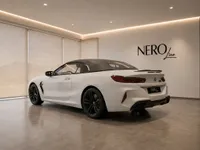 The white BMW M8 Competition Cabriolet, with its black soft top up, is parked in a showroom in Lausanne, viewed from the rear three-quarter angle.