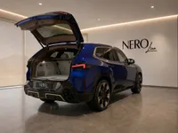 The blue 2023 BMW XM performance SUV is shown from the rear with its trunk open in a modern showroom in Lausanne.
