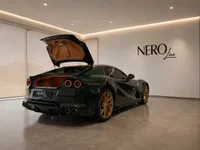 A dark green Ferrari 812 GTS with gold wheels is parked in a modern Zurich showroom, its trunk open revealing the interior.