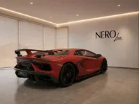 A matte red Lamborghini Aventador SVJ is showcased from a rear three-quarter view in a modern showroom in Lausanne.