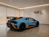 The light blue and orange Lamborghini Huracán STO is parked indoors, showcasing its dynamic rear three-quarter view in Lausanne.