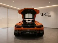 The orange Lamborghini Revuelto is shown from the rear with its engine bay open, inside a modern showroom in Lausanne.