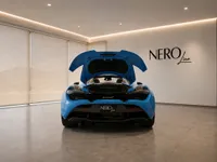 The blue McLaren 765LT Spider is shown from the rear with its engine bay open, parked in a showroom in Lausanne.