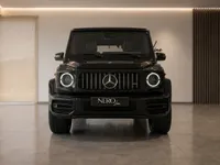 The front view of a glossy obsidian black Mercedes-AMG G 63 with its distinctive round headlights illuminated, parked in a modern showroom in Zurich.