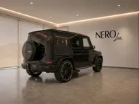 The gloss black Mercedes-Benz G63 Brabus is displayed from the rear three-quarter angle inside a modern showroom in Lausanne.
