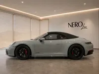 The gloss light grey Porsche 911 Carrera 4 GTS Cabriolet is displayed in a modern showroom in Zurich, showcasing its elegant side profile.