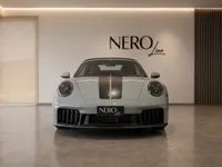 The gloss light grey Porsche 911 Carrera 4 GTS Cabriolet with a black racing stripe is prominently displayed in a modern NERO Line showroom in Zurich.