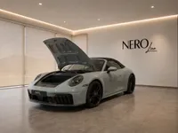 A gloss light grey Porsche 911 Carrera 4 GTS Cabriolet is displayed from the front with its hood open in a modern showroom in Zurich.