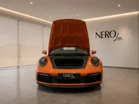 The orange Porsche 911 Carrera 4S is shown from the front with its frunk open, displayed in a showroom in Lausanne.
