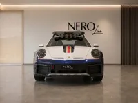 The Porsche 911 Dakar in gloss blue and white with gold and red stripes is shown from the front, featuring its roof rack, in a showroom in Zurich.