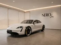 The white Porsche Taycan is showcased from a front three-quarter view in a modern NEROline showroom in Lausanne, with mountain views.