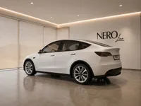 A white Tesla Model Y is parked indoors, viewed from the rear three-quarter angle in a modern showroom in Lausanne.