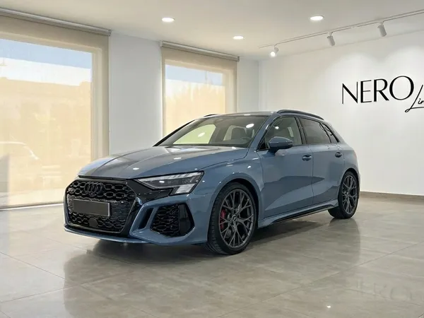 A sleek blue-gray Audi RS3 luxury sedan is showcased from a front three-quarter angle inside a bright showroom in Alicante.
