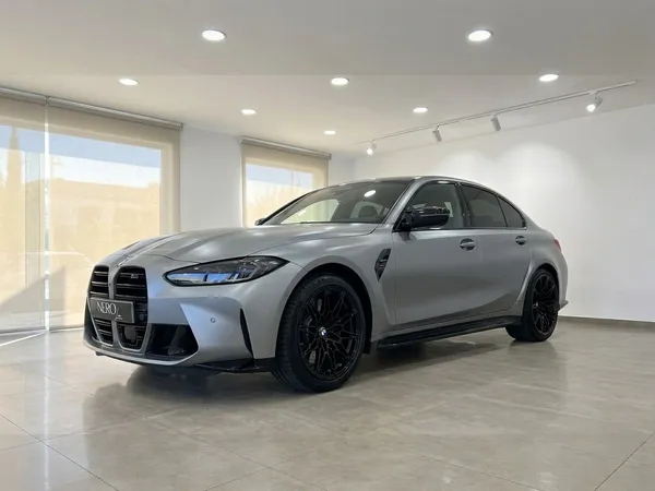 무광 프로즌 퓨어 그레이 BMW M3 컴페티션 xDrive가 마르베야 쇼룸에서 전면 쿼터 앵글로 돋보이게 전시되어 있습니다.