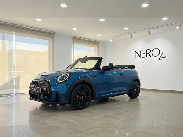 メタリックブルーのミニクーパー ジョンワークス コンバーチブルが、イビサのショールームでオープンルーフの状態で前方斜めから展示されています。