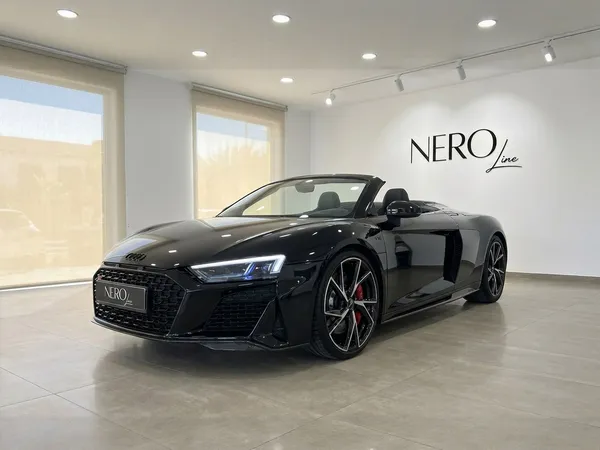 A gloss black Audi R8 Spyder convertible is parked in a bright showroom, viewed from the front three-quarter angle in Marbella.