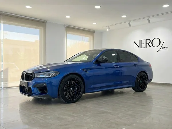 메탈릭 다크 블루 BMW M5 Competition Hybrid 럭셔리 세단이 마르베야 쇼룸에 앞측면에서 본 모습으로 주차되어 있습니다.