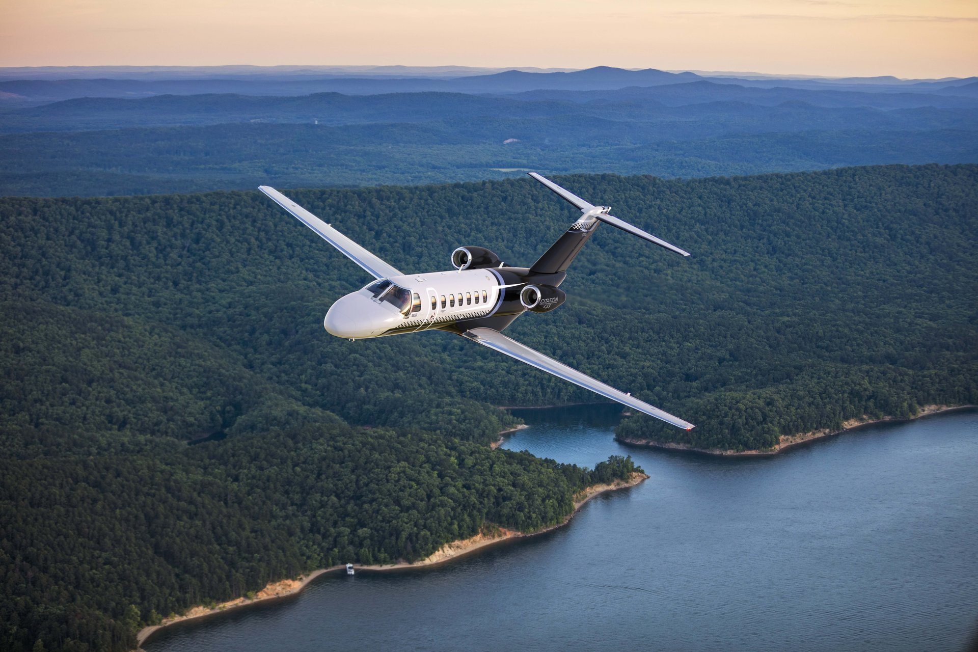 Cessna Citation CJ3 — In-flight over scenic landscape The sleek Cessna Citation CJ3 private jet soars gracefully over a lush green forested landscape with a winding blue lake below.