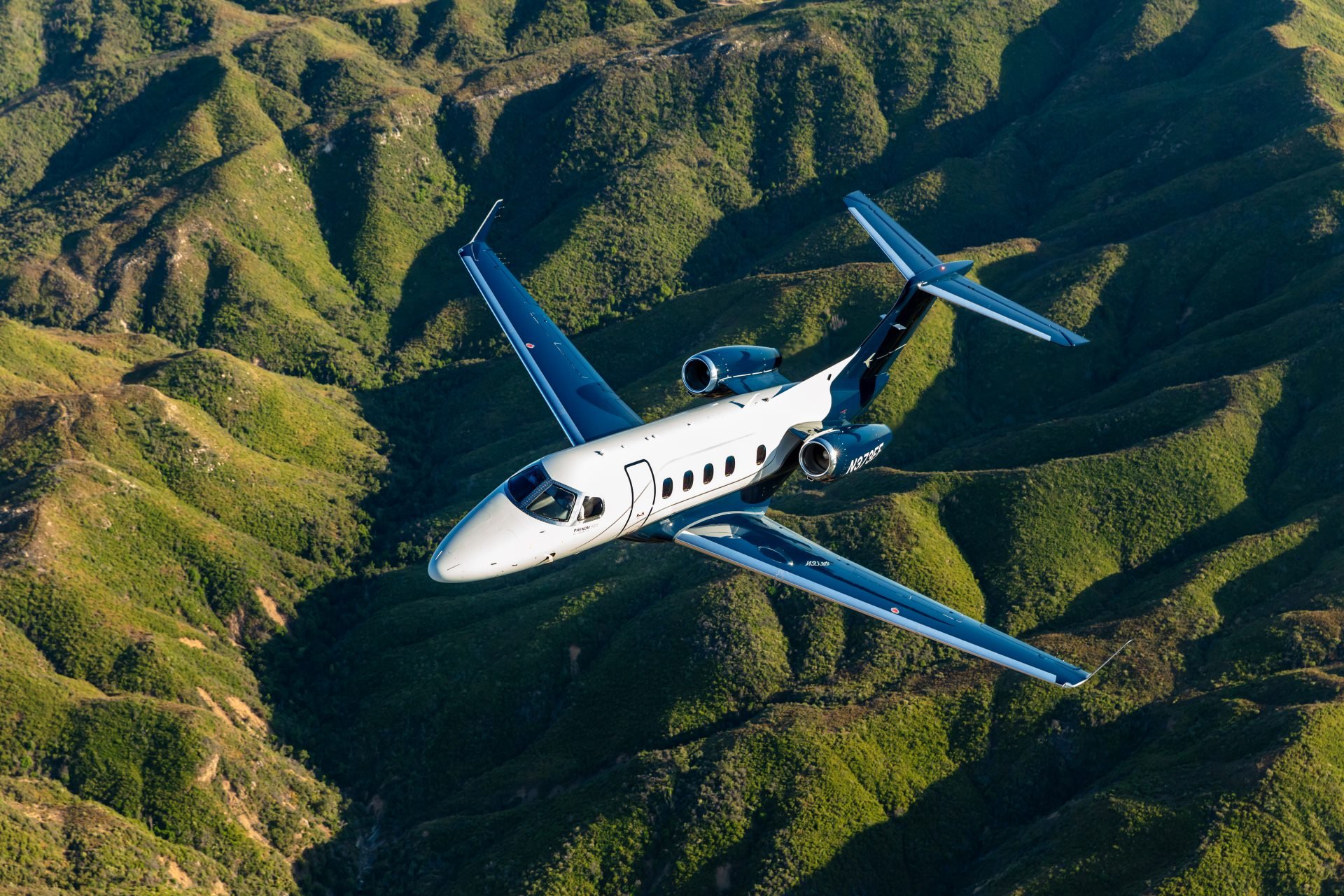 Embraer Phenom 300E — Flying over mountains The Embraer Phenom 300E light jet soars gracefully above lush green mountain ranges near Barcelona.