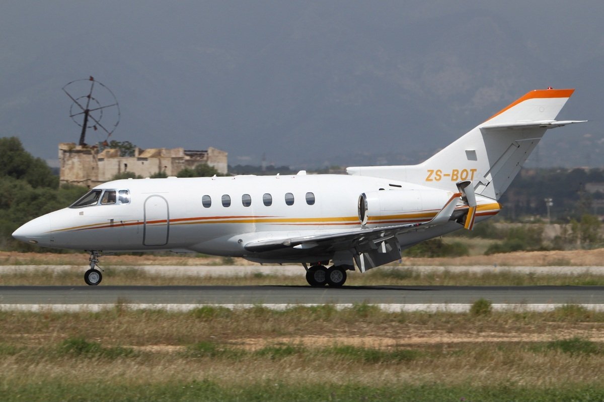 The Hawker 900XP private jet is seen from the side, taxiing on a runway in Malaga with historical buildings in the background.