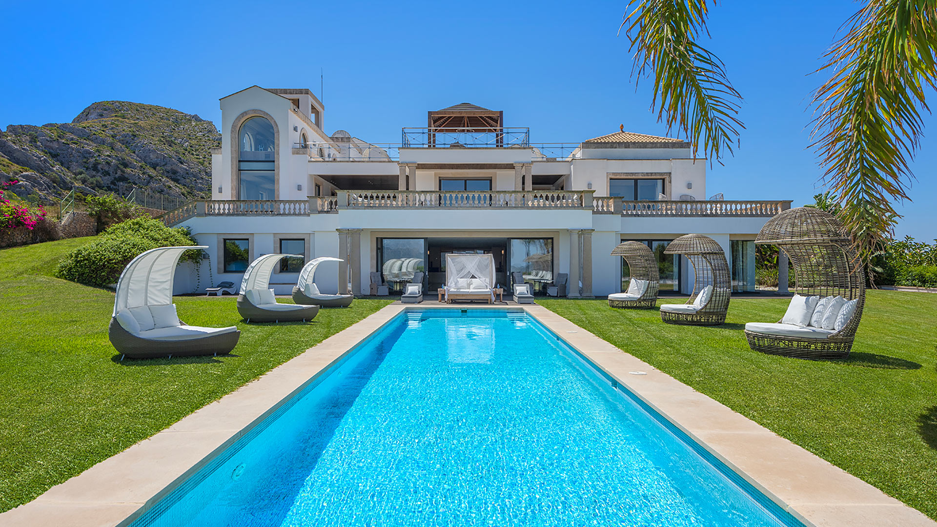 Villa Casa Ciel — Außenbereich mit Pool & Tagesbetten Die Villa Casa Ciel auf Mallorca mit einem langen Pool, üppigem grünen Rasen und stilvollen Tagesbetten im Freien, vor einer Bergkulisse.