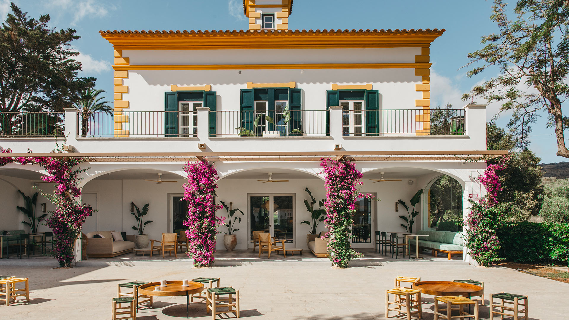 Hacienda Xini — Außenfassade und Innenhof Hacienda Xinis charmante weiß-gelbe Fassade mit grünen Fensterläden, einer Bougainvillea-Terrasse und Sitzgelegenheiten im Freien auf Menorca.