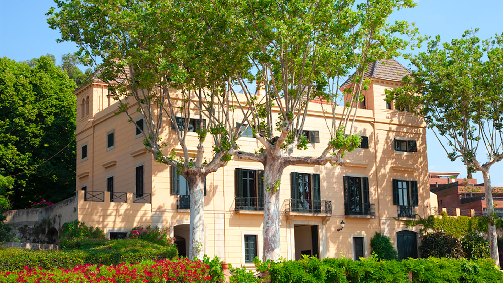 Villa Llorenc — Elegante Fassade und Garten Die elegante gelbe Fassade der Villa Llorenc wird von üppigen grünen Bäumen und leuchtend roten Blumen in ihrem Garten in Barcelona umrahmt.