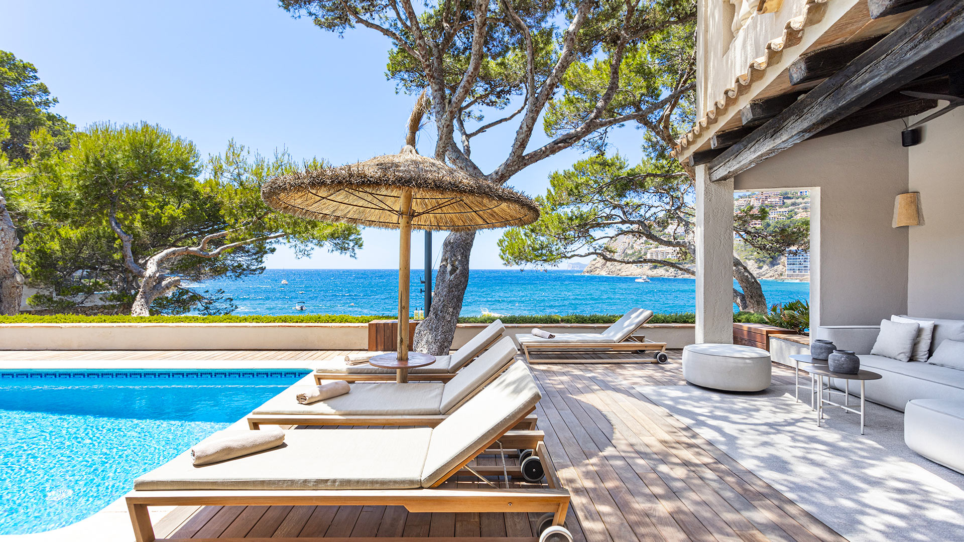 Villa Mañana — Pool-Lounge mit Meerblick Der Pool-Loungebereich der Villa Mañana mit Sonnenliegen, Strohschirm und dem Mittelmeer auf Mallorca.