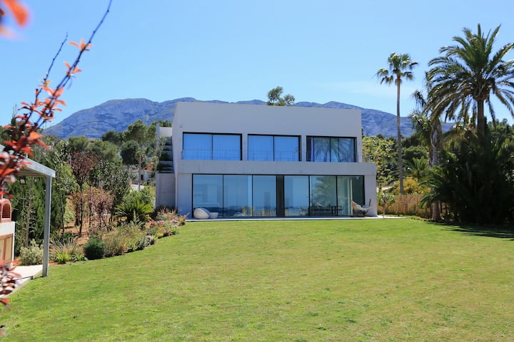 Villa Sea you Later — Moderner Außenbereich und Garten Moderner Außenbereich der Villa Sea you Later mit großen Glasfenstern, weitläufigem grünen Rasen und fernen Bergen in Denia, Spanien.