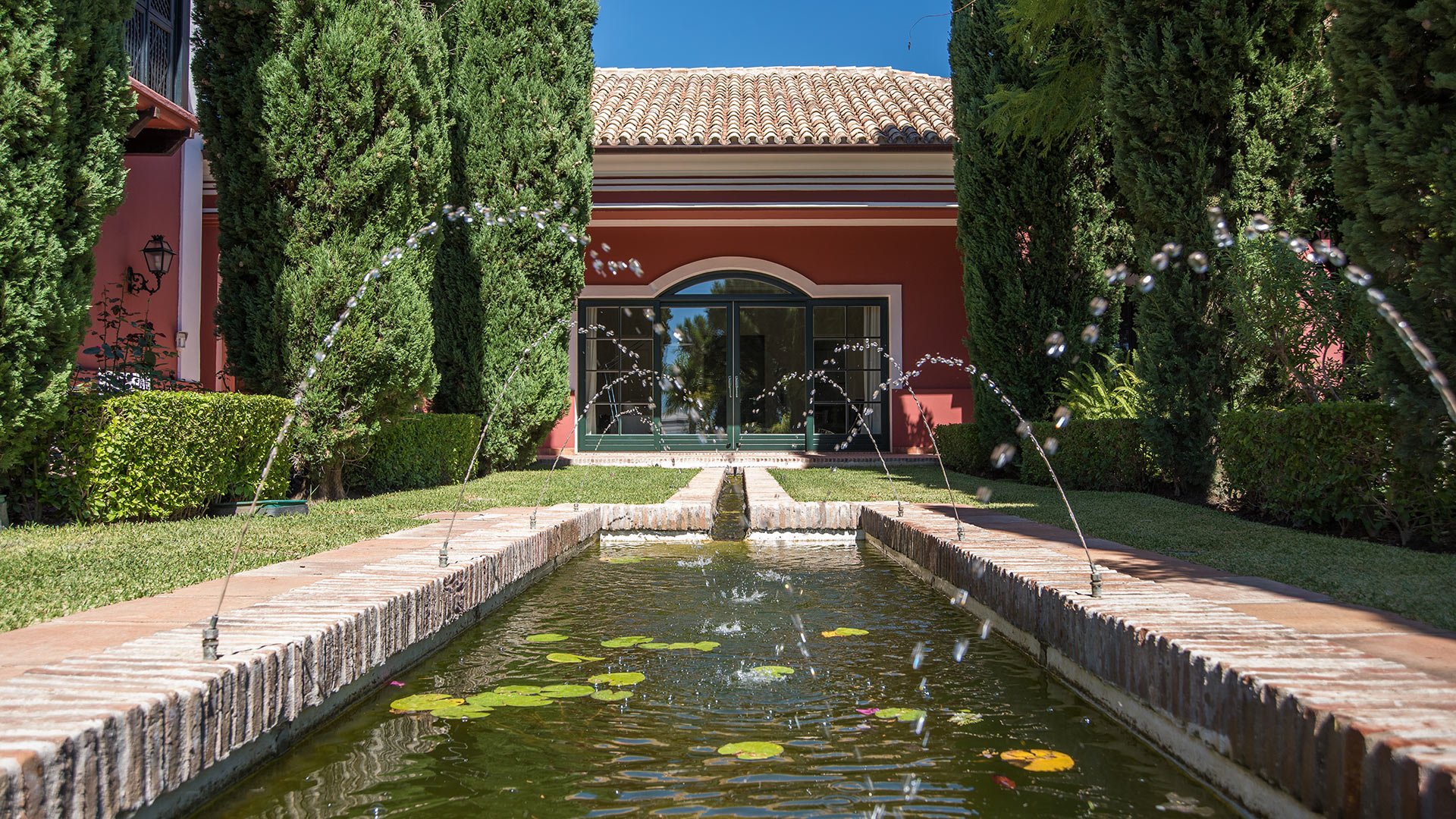 Villa Sombrera's elegant garden features a long brick fountain with water jets and lily pads, leading to the red villa in Marbella.