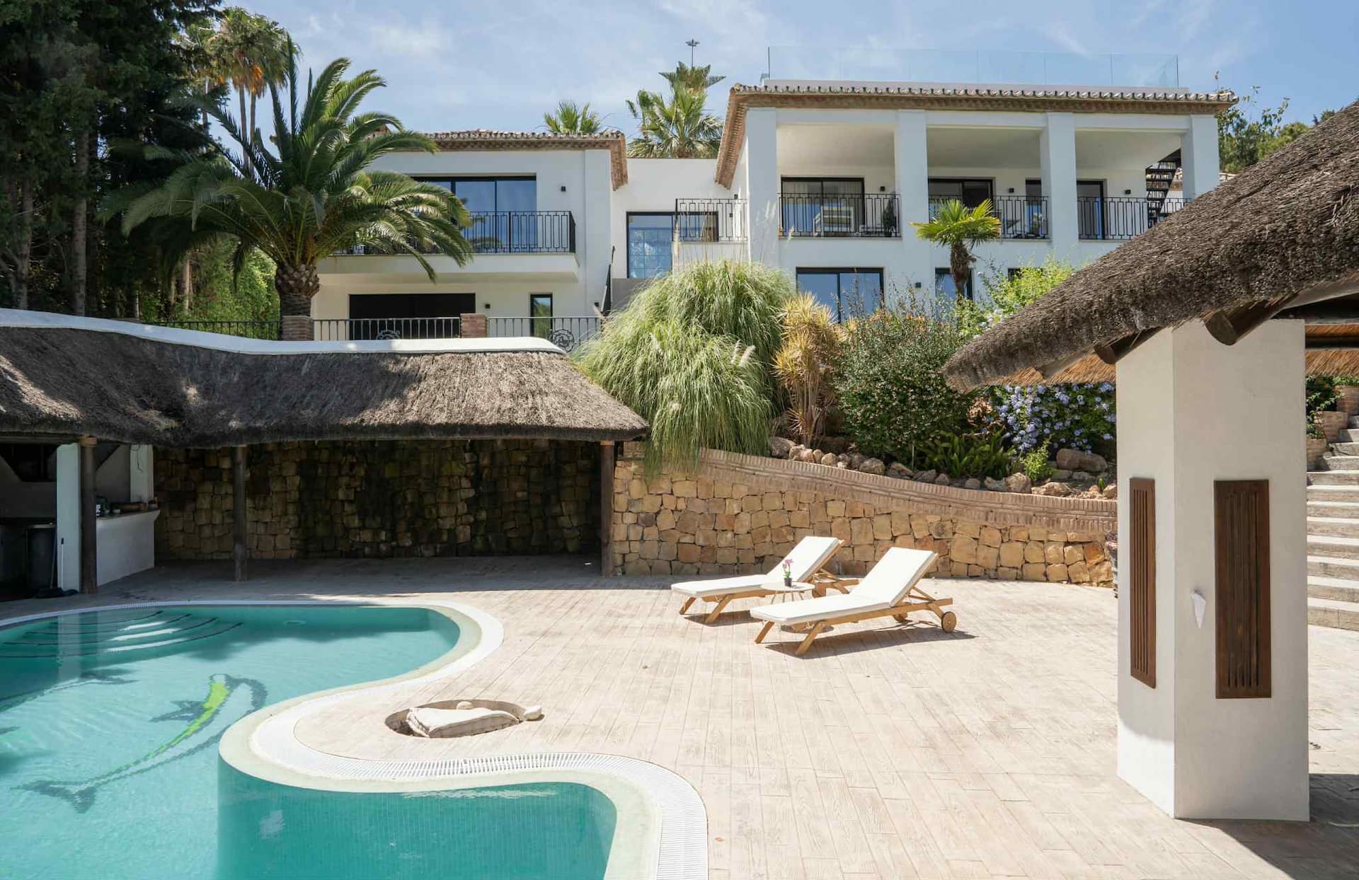 Villa Stella — Poolbereich & Außenküche Der Außenbereich der Villa Stella in Marbella zeigt einen geschwungenen Pool, Sonnenliegen, eine Bar mit Strohdach und das weiße Äußere der Villa.