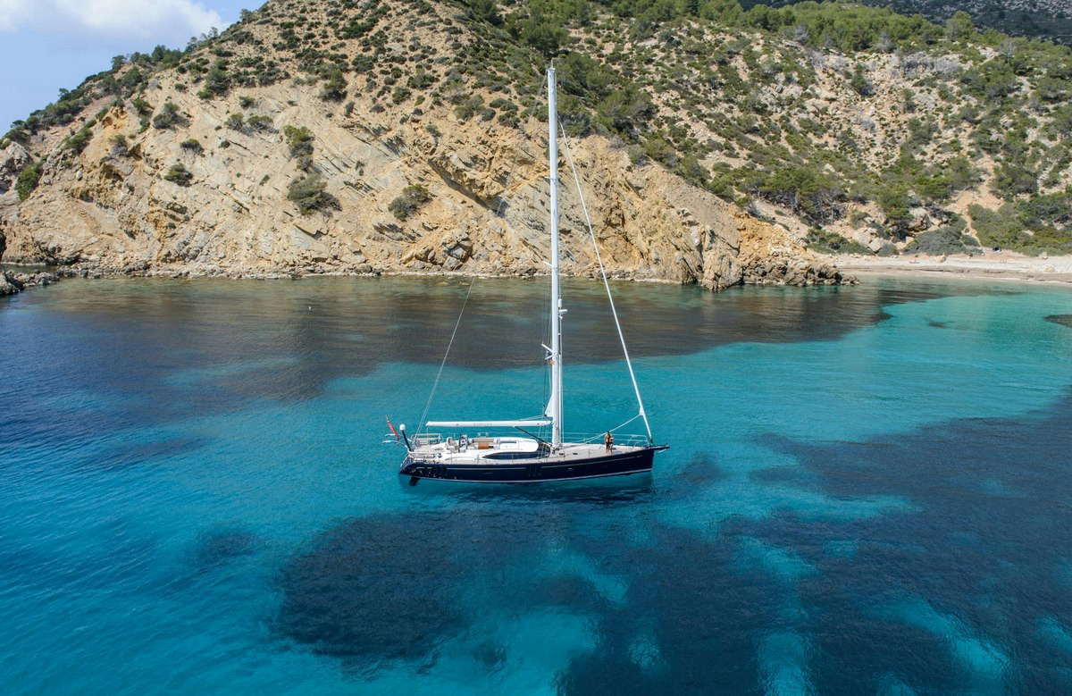 Oyster 675 — Anchored in a beautiful Ibiza cove The Oyster 675 sailing yacht is anchored in a serene turquoise cove near a rocky, vegetated coastline in Ibiza.