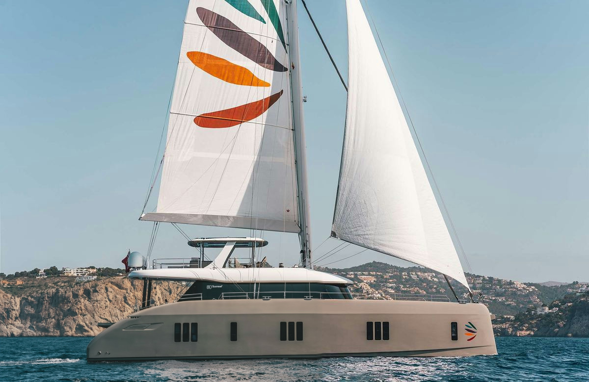 Sunreef 80 — Sailing catamaran off Ibiza The Sunreef 80 sailing catamaran glides on blue waters with full sails, one featuring colorful stripes, off the coast of Ibiza.