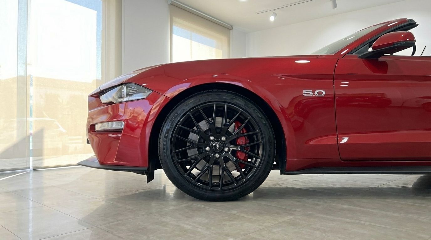 The racing red Ford Mustang GT Cabrio is shown from its front-side profile, highlighting the black multi-spoke wheel with red calipers in an Alicante showroom.